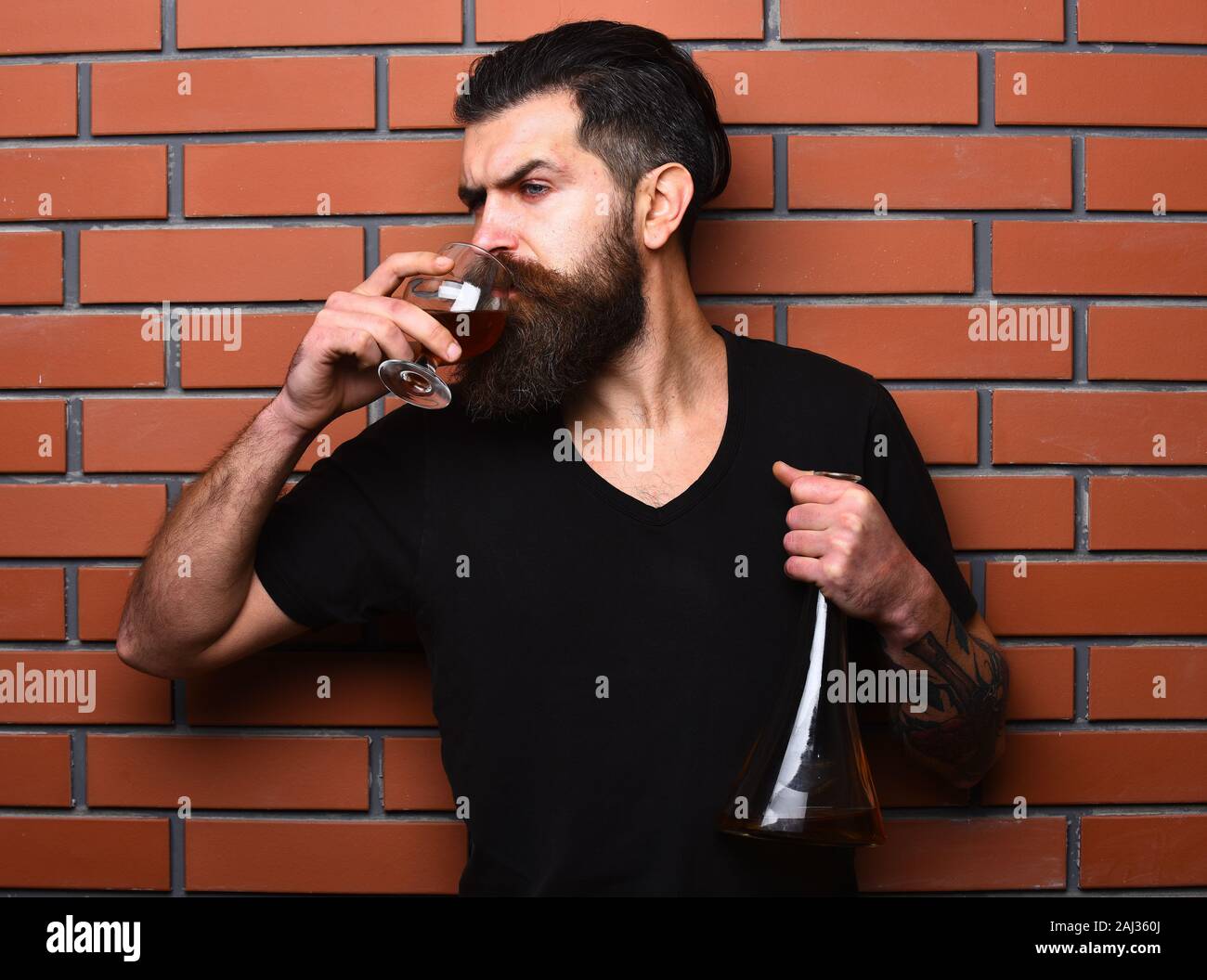 Guy with glass and bulb of cognac. Sommelier with busy face drinks brandy or whiskey. Service and tasting concept. Man with beard and mustache holds alcoholic beverage on brick wall background. Stock Photo