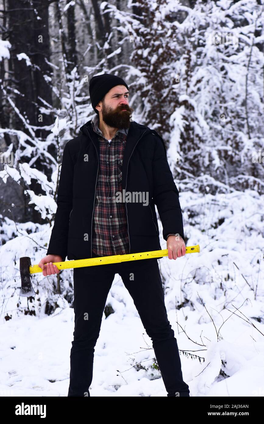 Winter hiking concept. Man in hat with beard and mustache holds ax. Guy with thoughtful face with forest covered by snow on background. Forestman with yellow axe Stock Photo