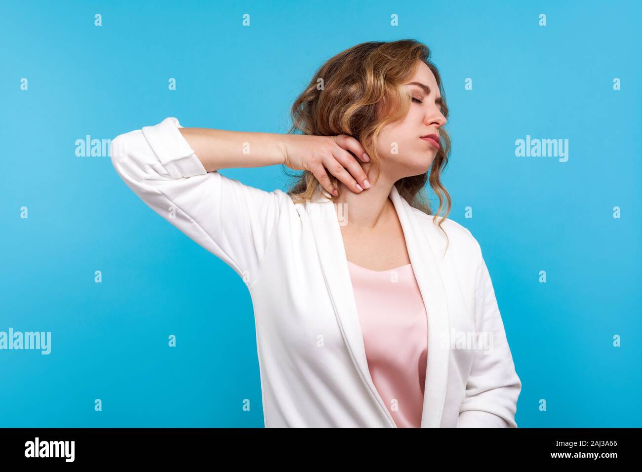 Portrait of tired woman with wavy hair in white jacket suffering neck ache, standing with closed eyes and massaging neck to relieve pain, feeling exha Stock Photo
