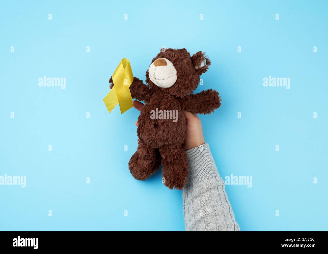 female hand hold a small brown teddy bear which holds in its paw a yellow ribbon folded in a loop on a blue background. concept of the fight against c Stock Photo