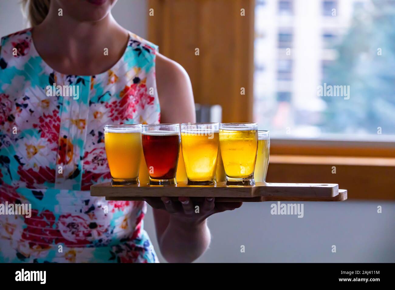 selective focus on Flight of Craft four of different Beers glasses on Wooden two Tray, a caucasian woman serving holding the trays in the background Stock Photo