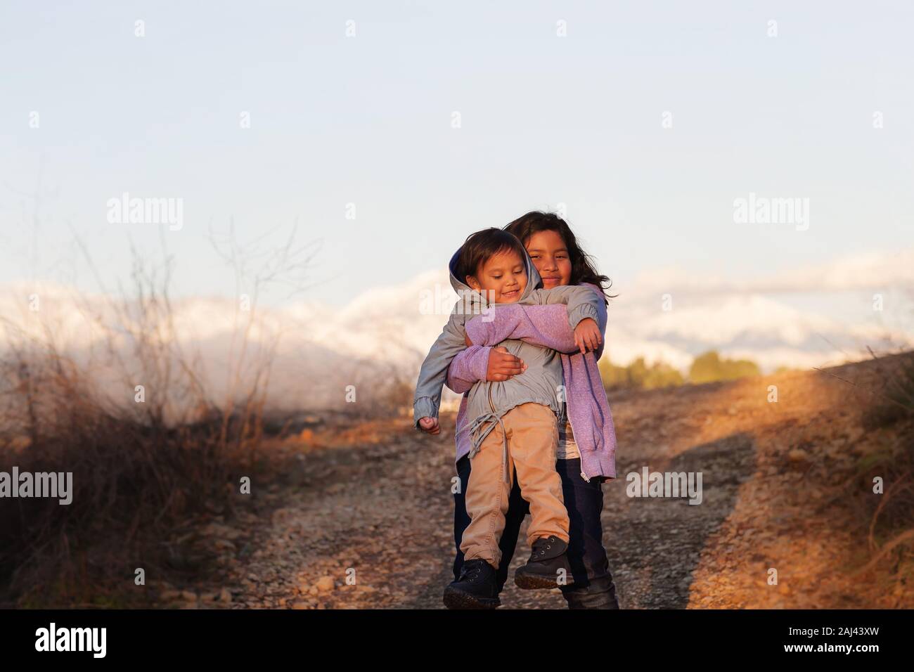 An older sister hugging and holding up her little brother while standing in the middle of a hiking trail with mountains in the background. Stock Photo