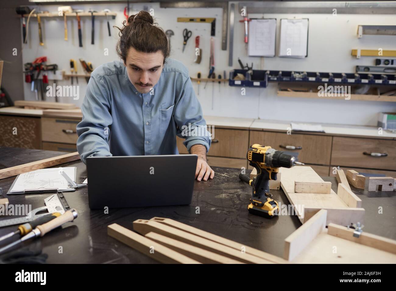 Young carpenter using laptop while standing at the table with wooden ...