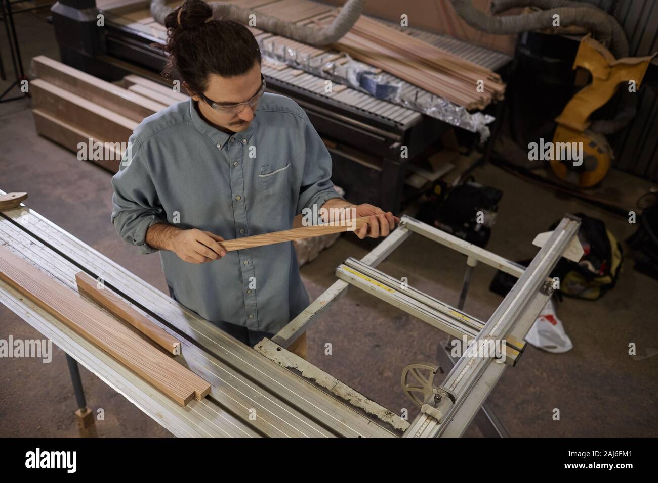 Young carpenter examining the wooden plank in his hands while working on lathe in workshop Stock Photo