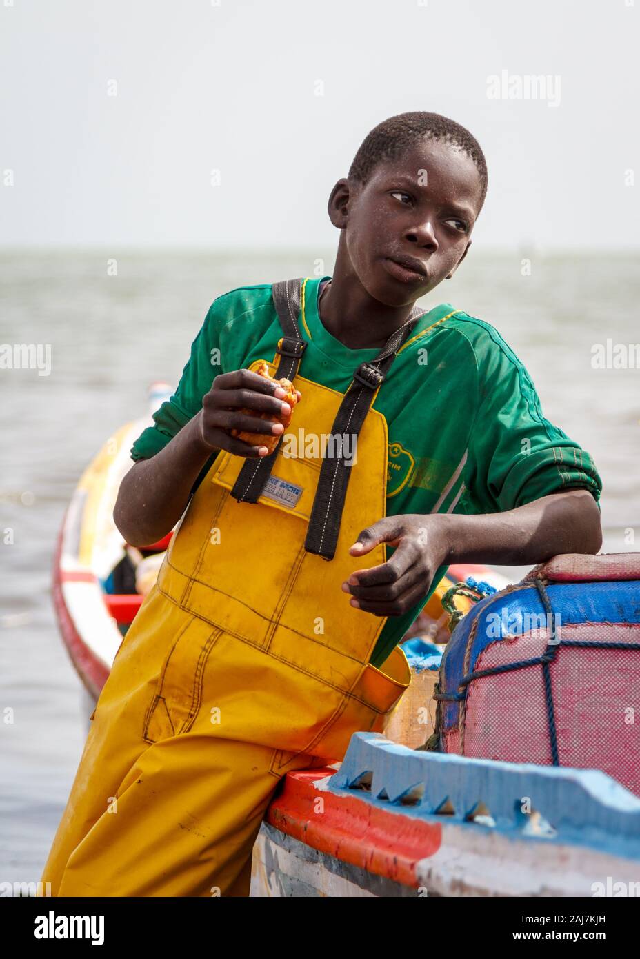 Portrait of a young fisherman casually leaning against his colourful fishing boat. Fishing Port in Joal Fadiout, Senegal, Africa - Photograph: Tony Ta Stock Photo