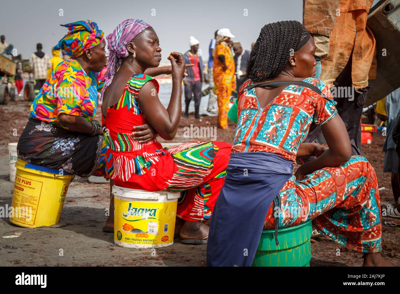 Three women colourfully dressed waiting for the catch on the beach on the coast by Fishing Port in Joal Fadiout, Senegal, Africa - Photograph: Tony Ta Stock Photo