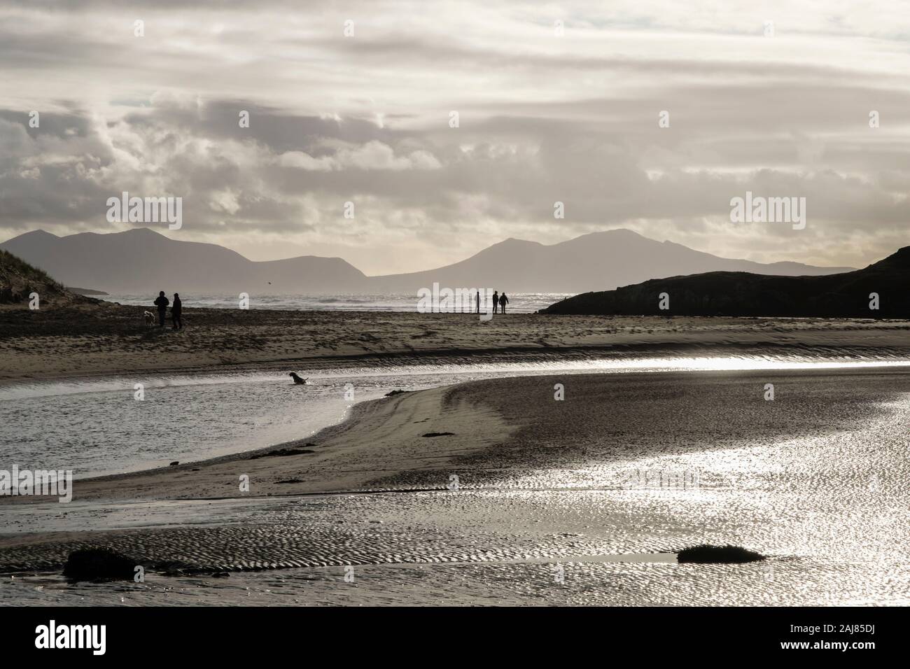 Backlit view across Afon Ffraw river with people walking on Traeth Mawr beach with mountains beyond in winter. Aberffraw Isle of Anglesey Wales UK Stock Photo