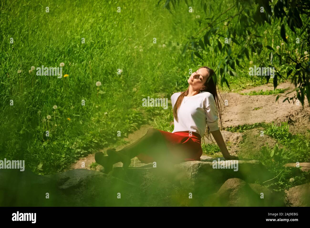 Portrait of a girl in red skirt and white blouse sitting on stone on river bank Stock Photo