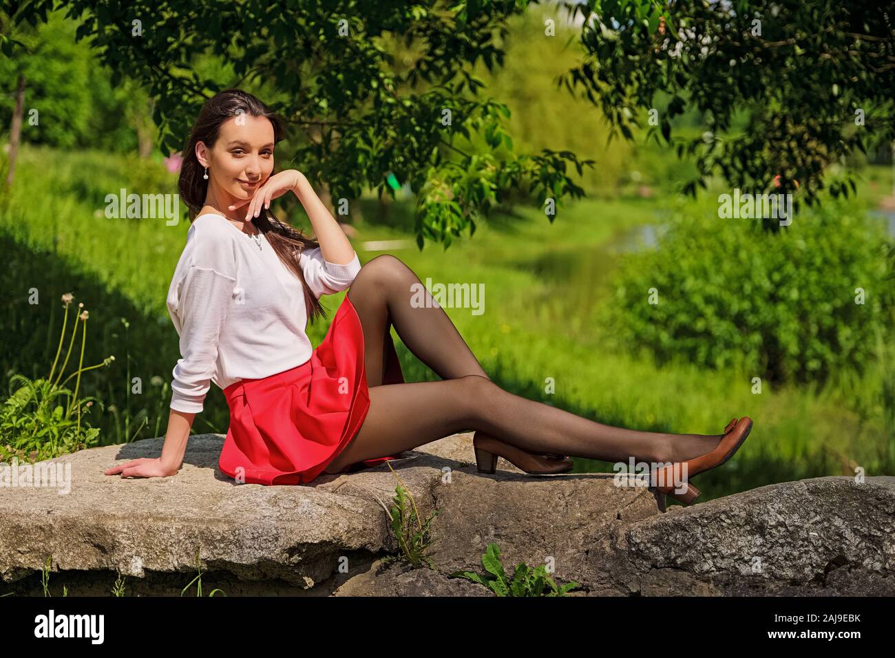 Portrait of a girl in red skirt and white blouse sitting on stone on river bank Stock Photo