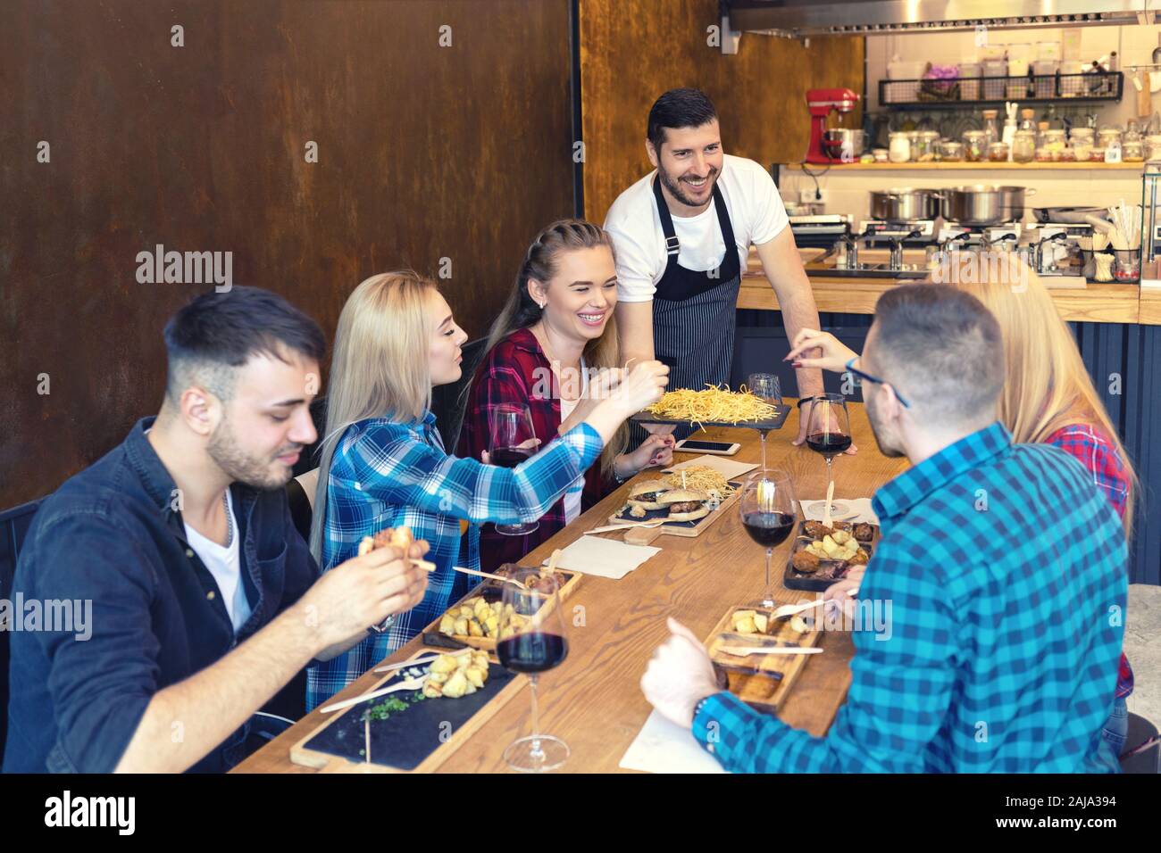 Happy business owner serving food to smiling group of young friends in small family tavern restaurant Stock Photo