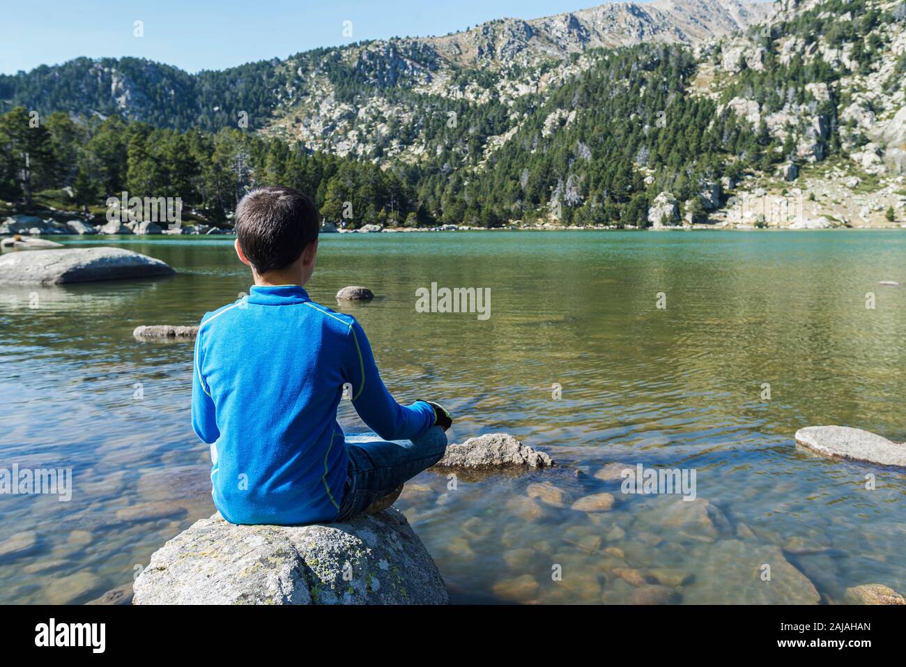 Rear view of a boy sitting on rock on a mountain lake looking away ...
