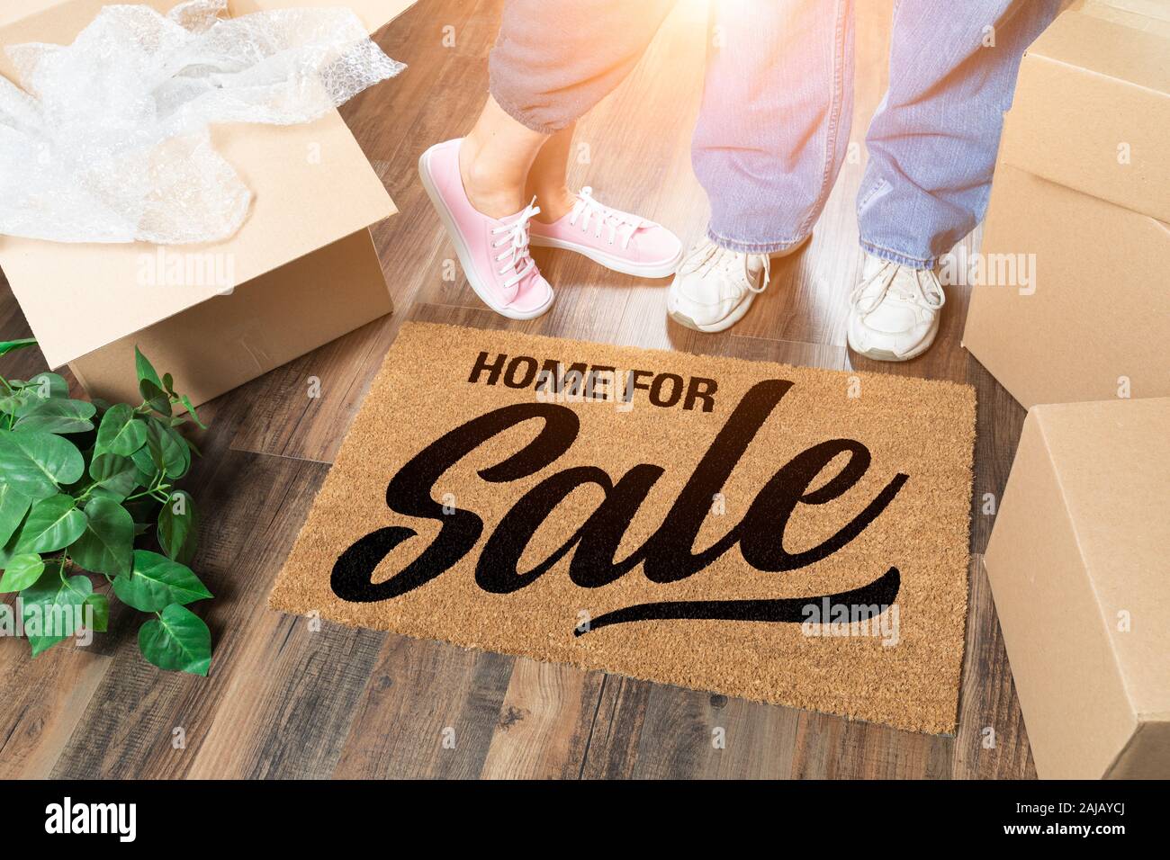 Man and Woman Standing Near Home For Sale Welcome Mat, Moving Boxes and Plant. Stock Photo