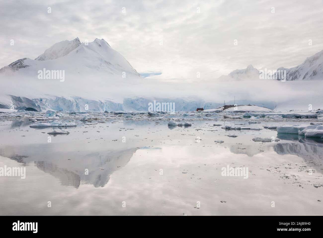 Port Lockroy (penguin post office) with mountains and glacier behind and ice and reflections in front, Antarctica Stock Photo