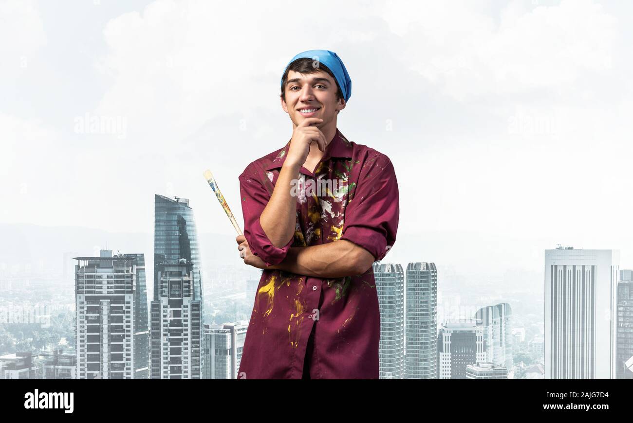 Smiling young painter artist holding paintbrush. Portrait of happy art school studentin shirt and bandana on cityscape background. Artistic occupation Stock Photo