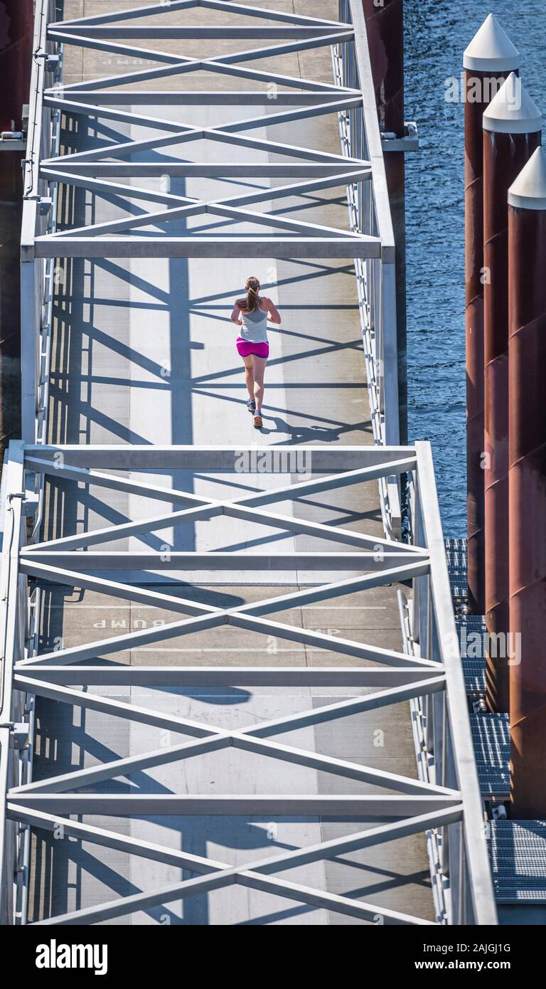 Woman in sportswear engaged in amateur sports running using a compact bridge connecting two parts of a floating dock on the river as a running shake, Stock Photo
