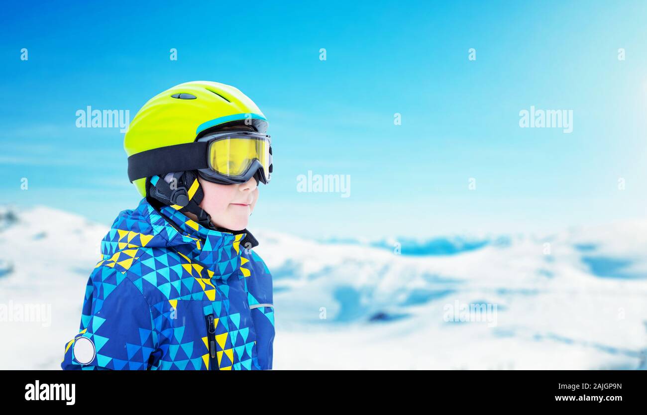 Boy with modern ski equipment at the top of a snowy mountain. Text space beside. Snowy peaks of the ski resort in background. Green helmet, yellow gla Stock Photo