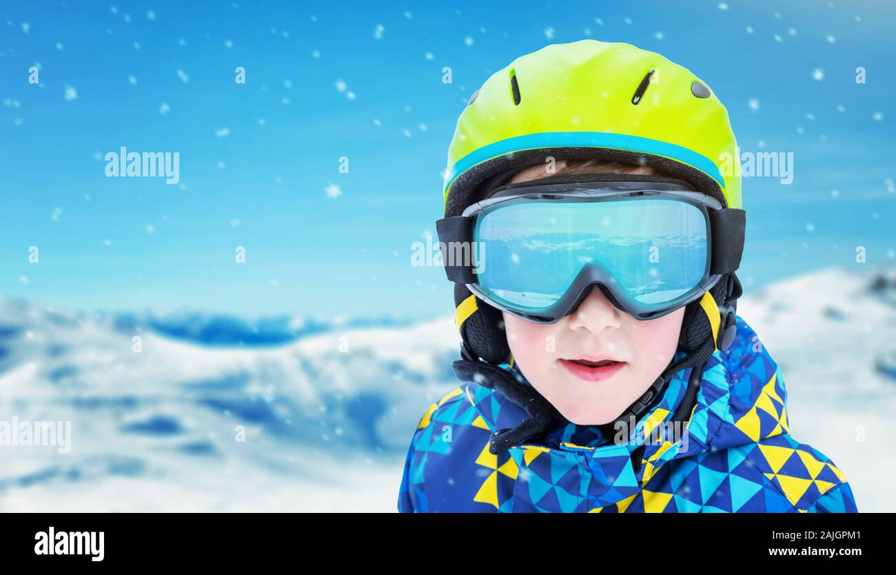 Boy with modern ski green helmet, yellow glasses and blue jacket. Reflection of snowy peaks on glasses. Close-up, copy space beside Stock Photo