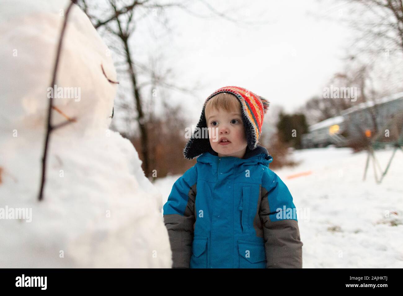 Mid shot of toddler boy wearing knit hat standing looking at snowman Stock Photo