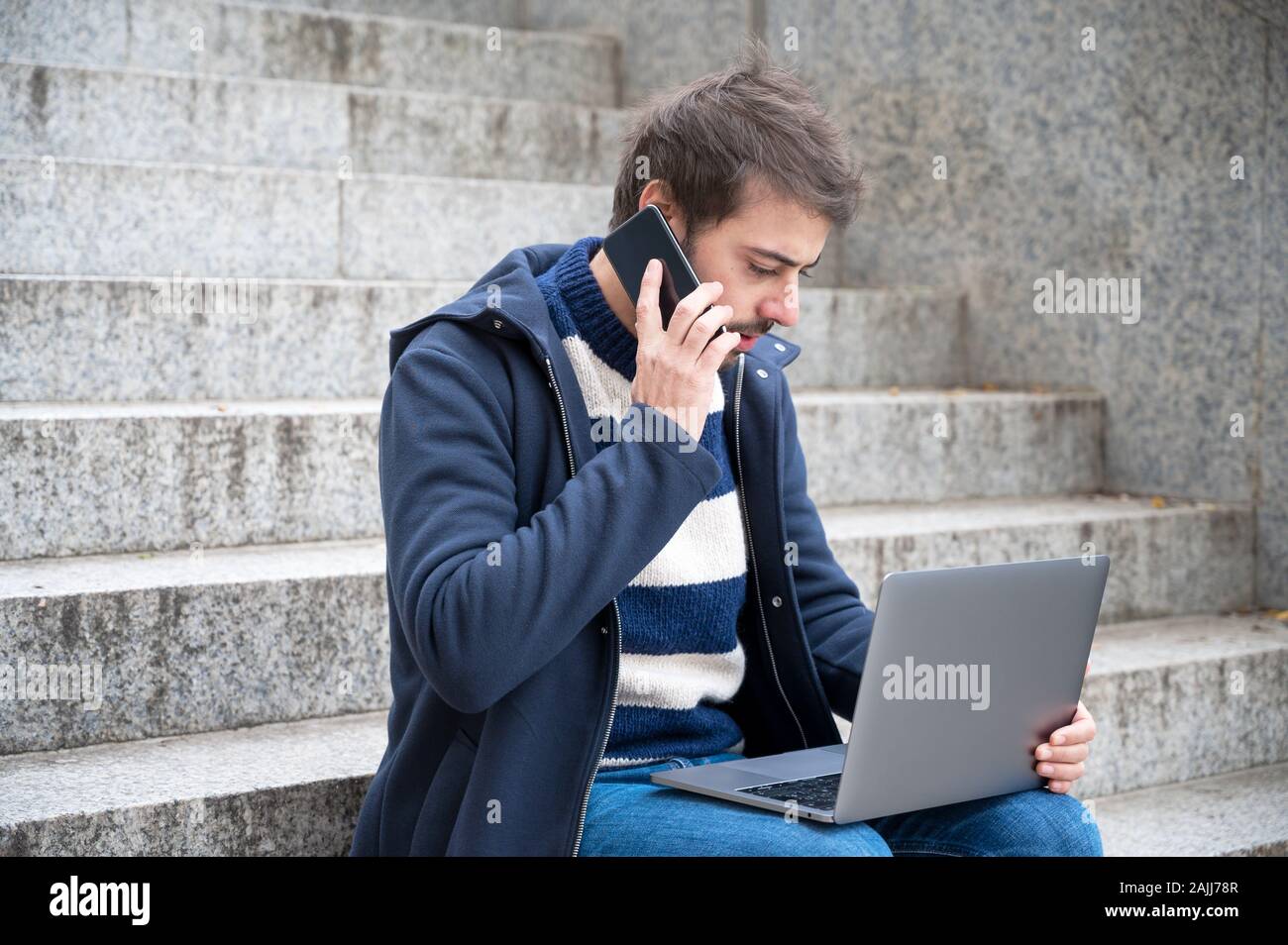 Man talking on phone while working on laptop sitting in stair steps in urban scenery . Stock Photo