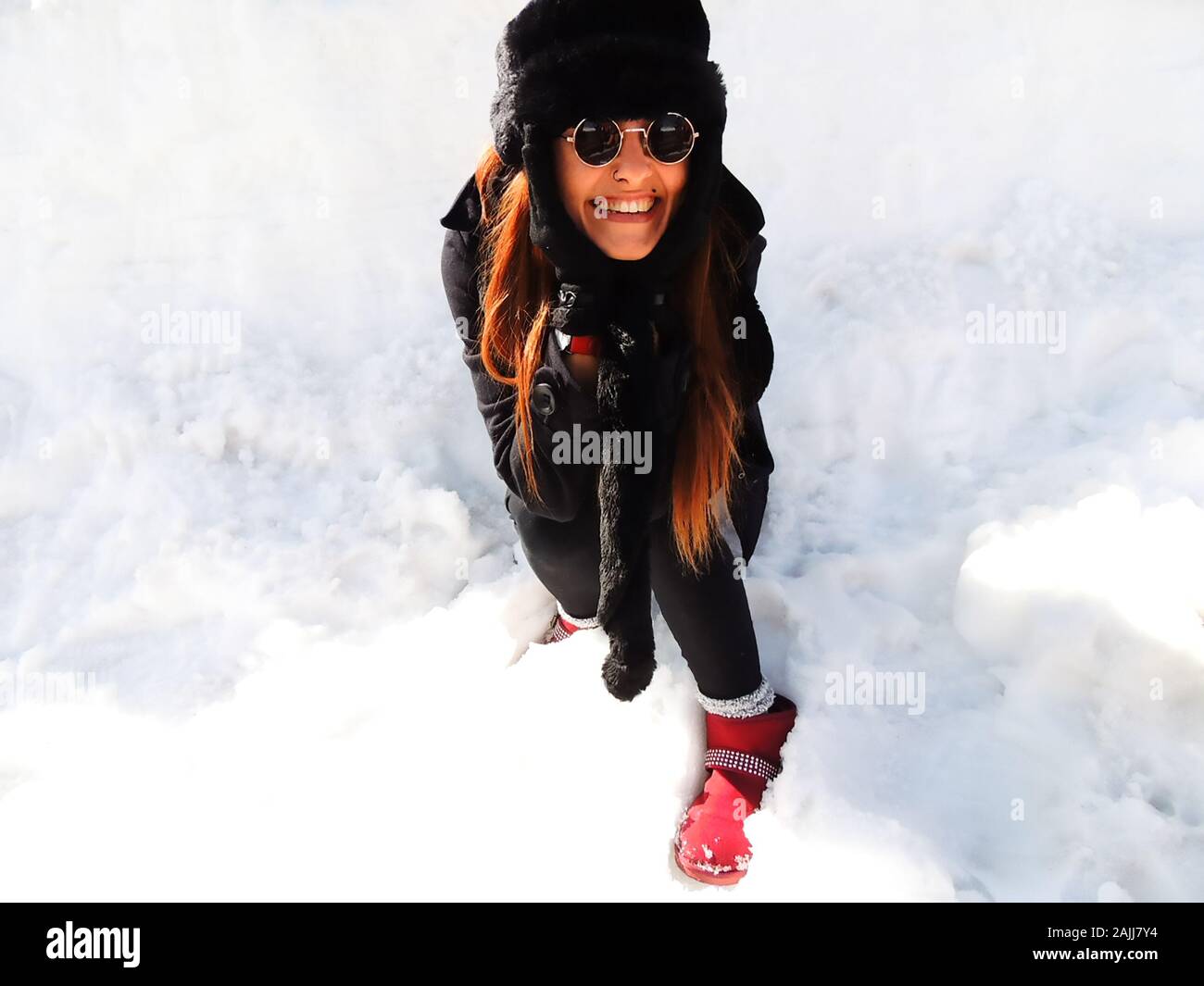 Stylish expressive redhead woman surrounded with snow wearing winter clothing, fluffy hat, fashion sunglasses and red shoes looking at the camera Stock Photo