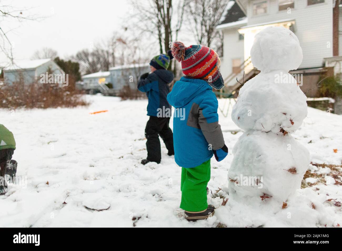 Preschool age boy stands next to snowman while brothers play in snow Stock Photo