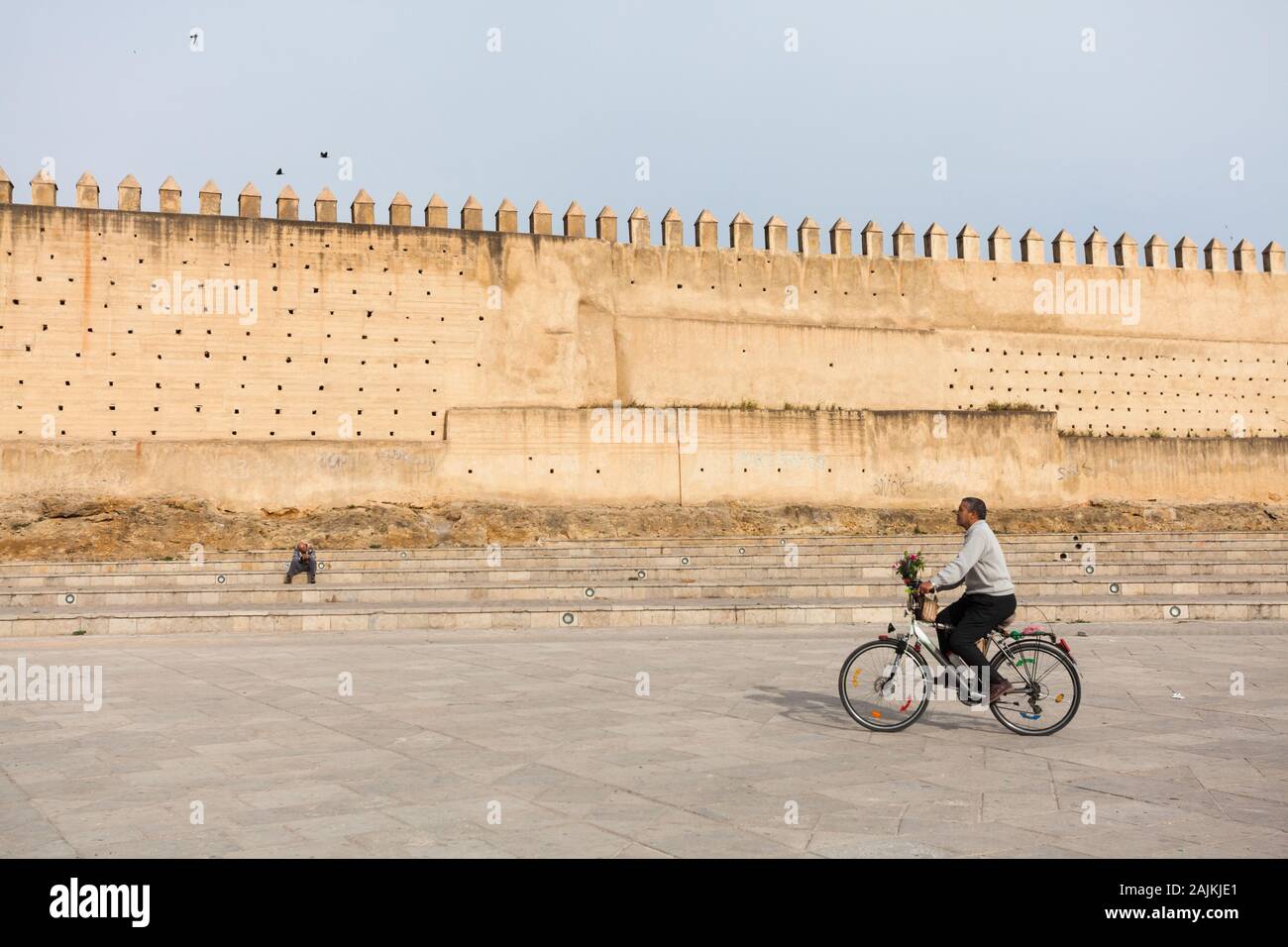 Man with a bunch of flowers riding a bicycle in the scenery of Boujloud Square (Place Boujloud) and the rammed earth city walls of Fes (Fez), Morocco Stock Photo