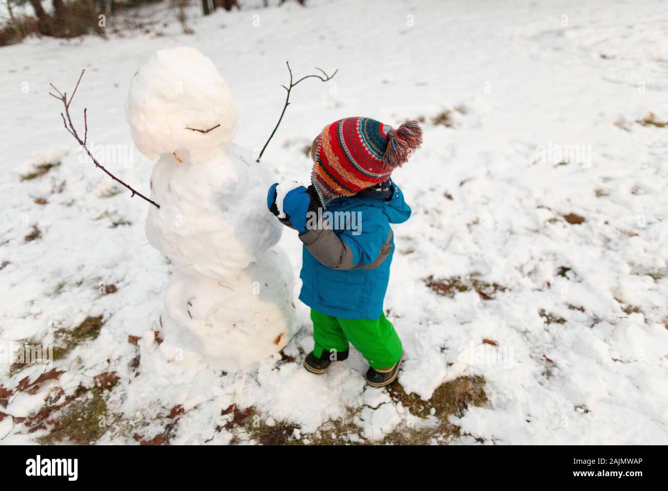 Toddler wearing cold weather gear stands next to snowman holding snow Stock Photo