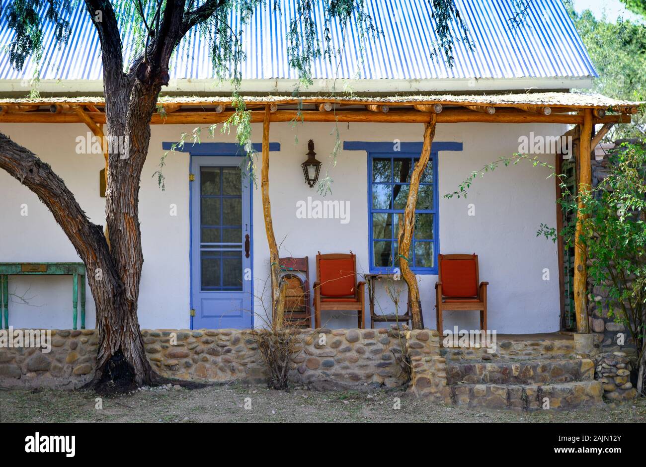 A Mesquite tree near the pole and tree post porch of a Spanish influenced adobe home with red chairs on porch in artisan town of Tubac, AZ, USA Stock Photo