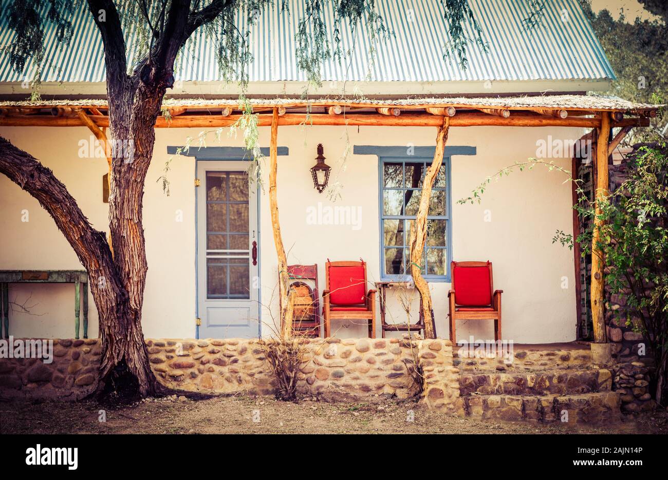 A Mesquite tree near the pole and tree post porch of a Spanish influenced adobe home with red chairs on porch in artisan town of Tubac, AZ, USA Stock Photo