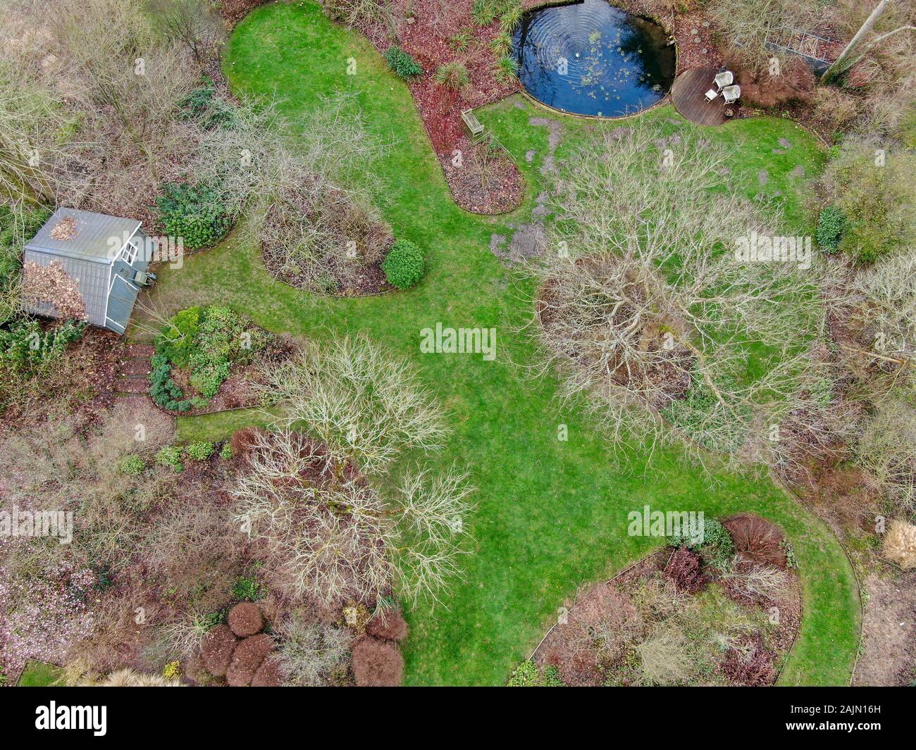 Aerial view of English garden type during winter season. Garden with a large green grass, little pound of water and different type of plants, trees and small garden wood house. Stock Photo