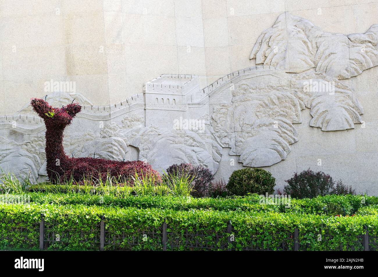At the Badaling site of the Great Wall of China, a sculpted shrub deer sits before a wall carving depicting one of the 7 Wonders of the World. Stock Photo