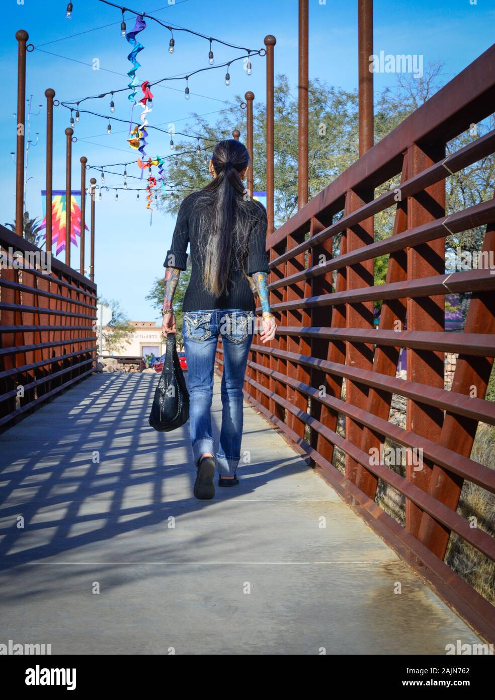 Rear view of hipster woman with tattoo sleeves strolling across the modern, metal, industrial designed pedestrian walkway in Tubac, AZ Stock Photo