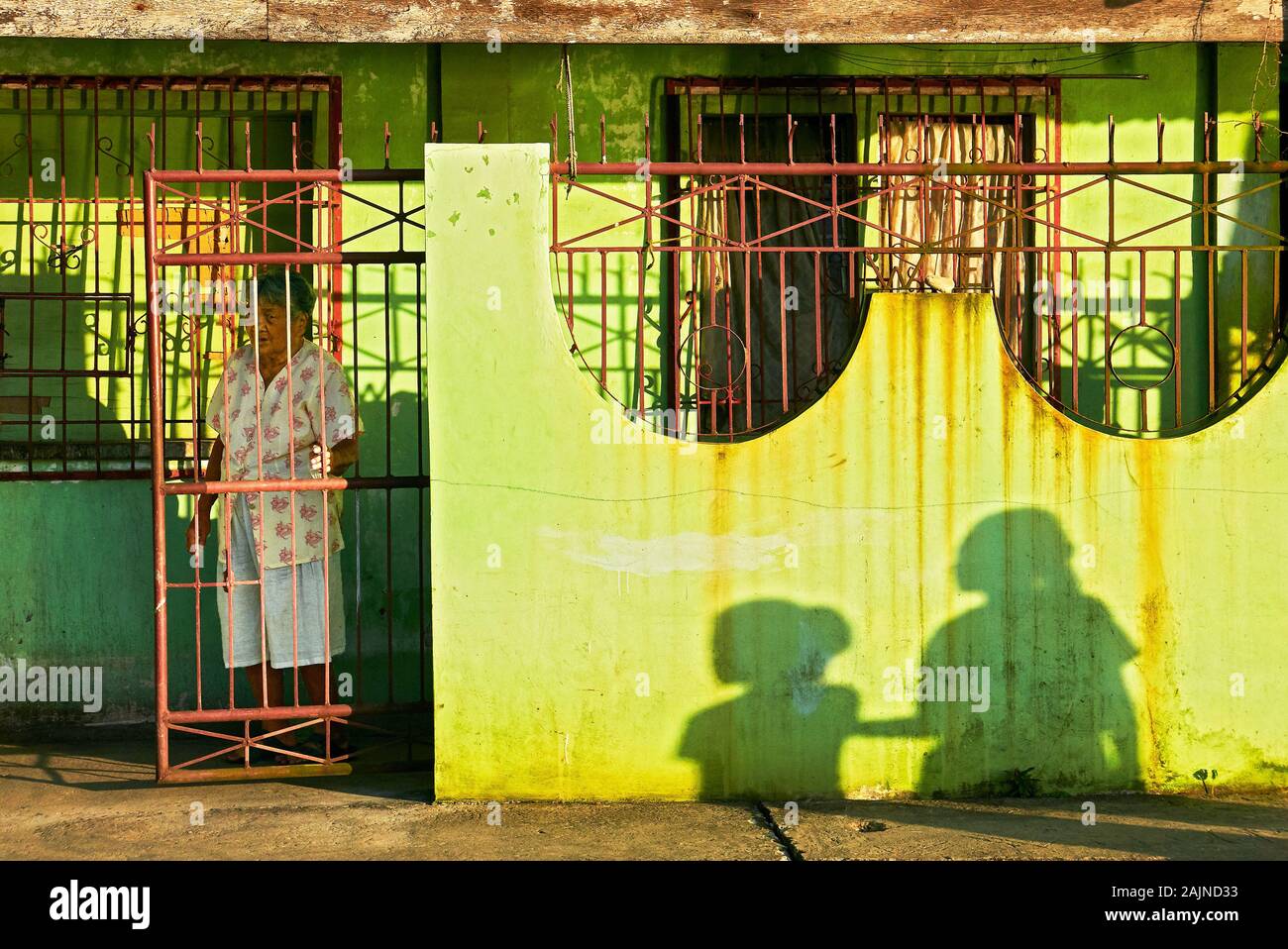 Romblon Town, Philippines: Poor green painted building with iron doors at late afternoon, old woman and shadows of two girls on wall Stock Photo