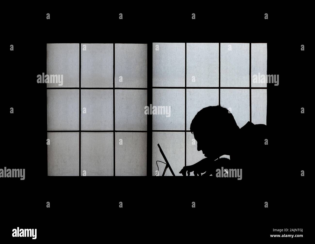 Window silhouette of a creepy computer hacker hunched over a laptop late at night Stock Photo