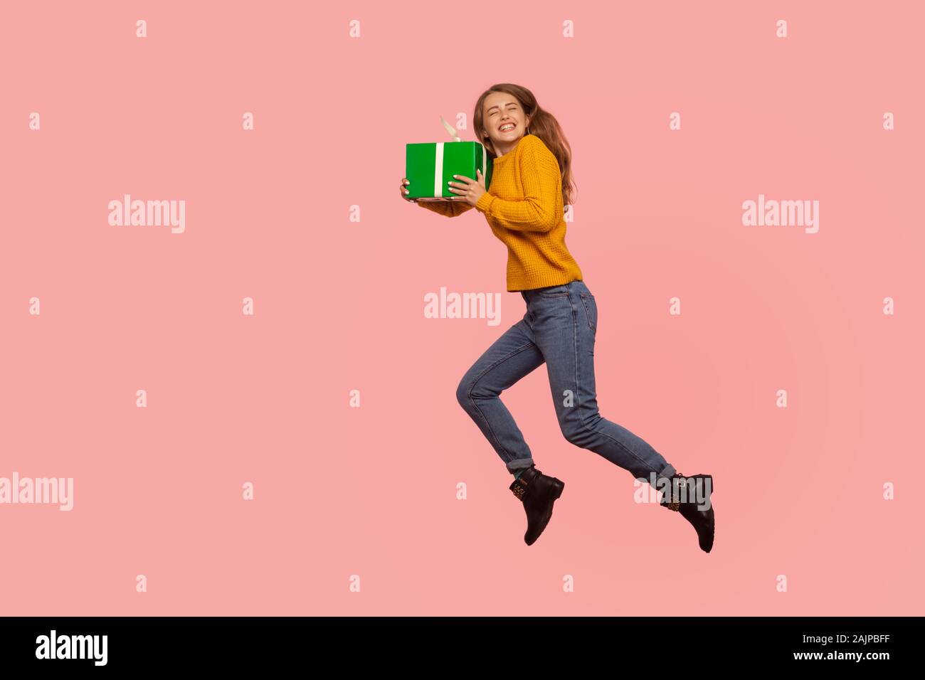 Portrait of extremely happy ginger girl in sweater and denim jumping in air with gift box, full of energy and enthusiasm flying with holiday present. Stock Photo
