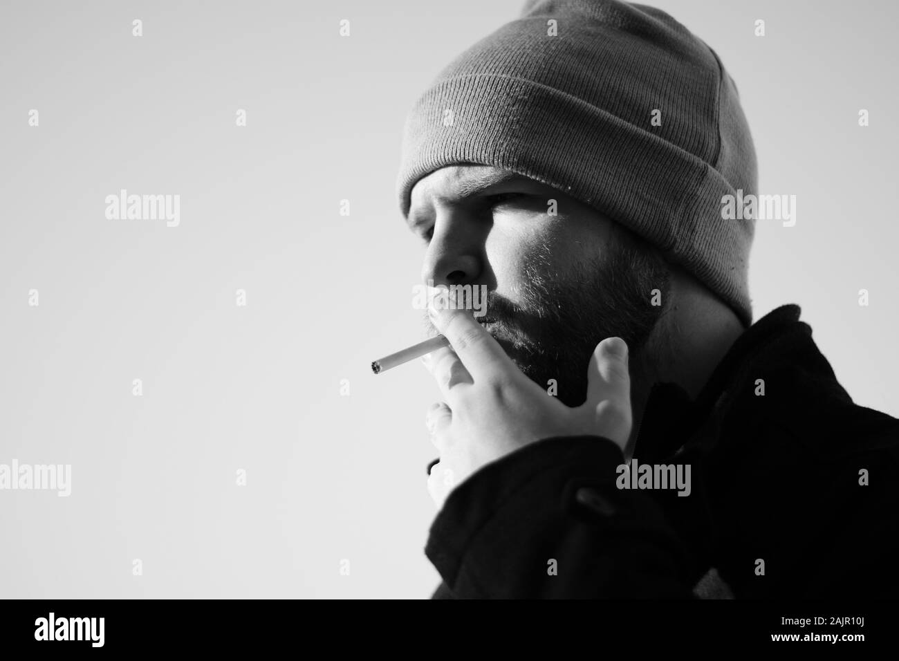 Caucasian bearded male with a beanie holding a cigarette, harshing sunlight  with strong shadows. Narrow shot black and white Stock Photo