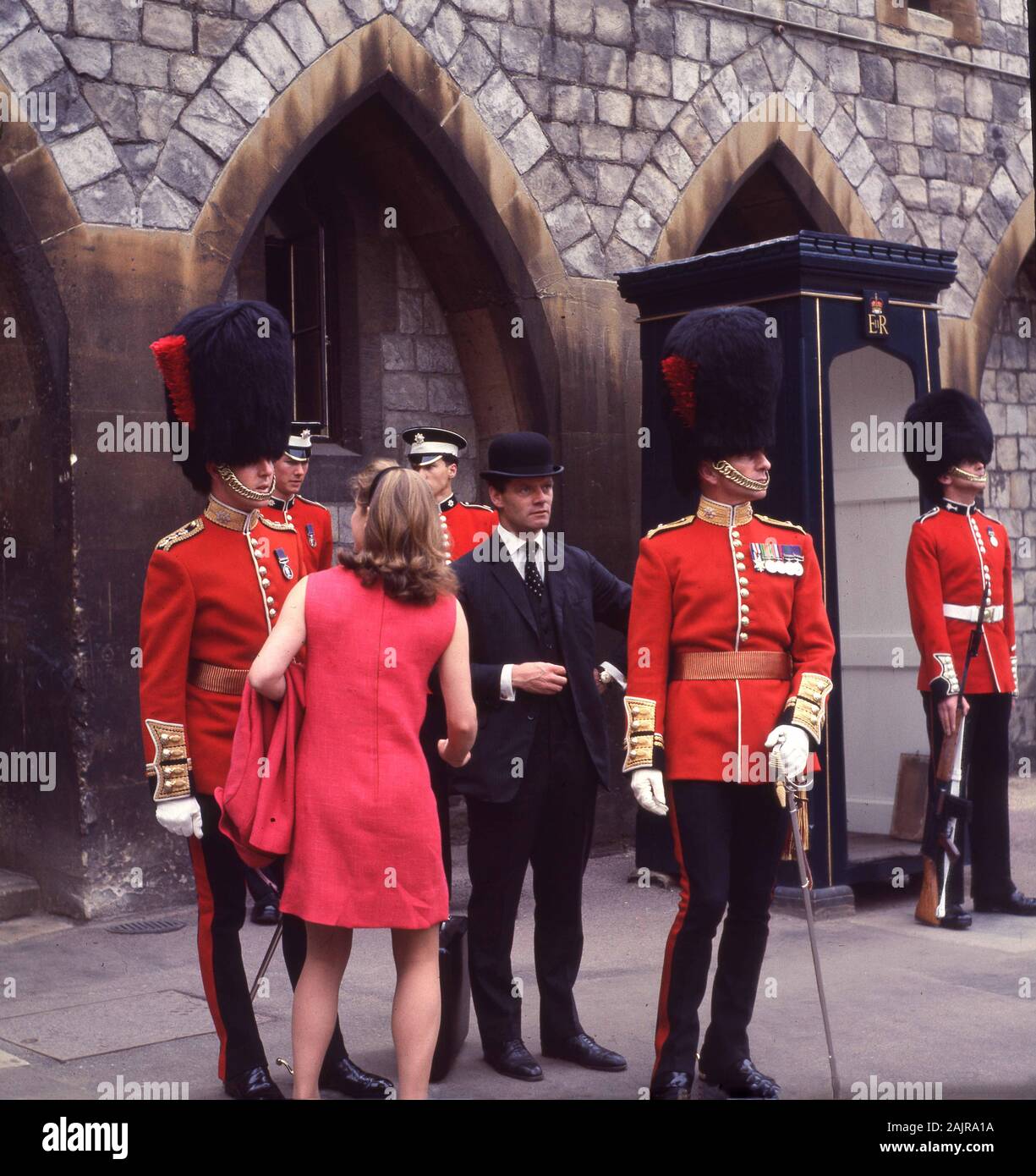 1960s, historical, at the most Noble Order of the Garter day at Windsor Castle, Windsor, England, a gentleman in a bowler hat and a lady in a red dress looking at the uniformed Queen's Guards wearing  their bearskins on ceremonial duty. The most Noble Order of the Garter was founded by King Edward III of England in 1348 and is the most senior order of Knighthood in the British honours system. Stock Photo