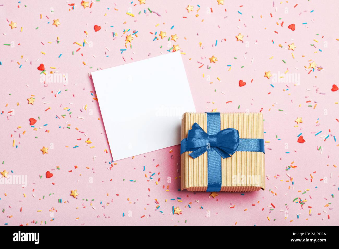 A gift in kraft box with blue bow and paper for notes on a pink background with decorations. Surprise your loved one. The concept of the day of St. Valentine's, birthday and other holidays. Flat lay Stock Photo