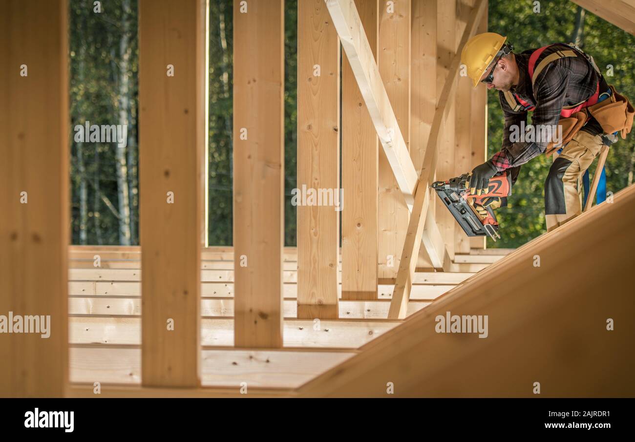 Construction Worker Contractor in His 30s With Nail Gun Attaching Wooden Elements. Wearing Hard Hat and the Wooden House Frame. Stock Photo