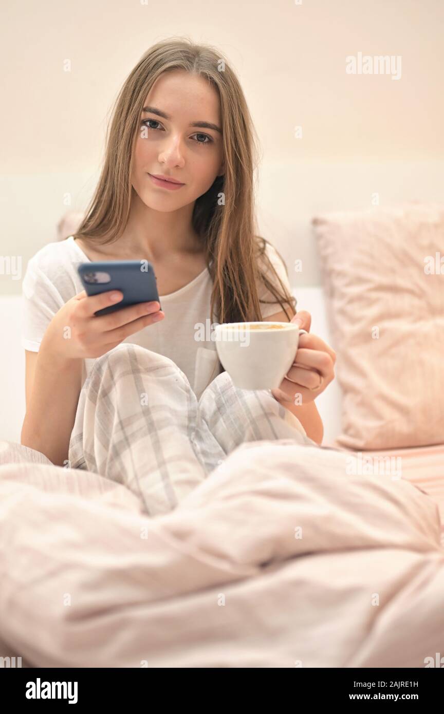 Cheerful Young Woman Sitting In Bed With Smartphone And Cup of Coffee Stock Photo