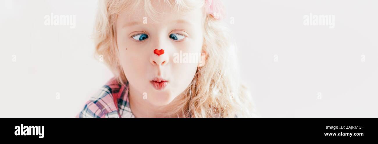 Cross-eyed kid looking at red heart sticker on nose. Funny hilarious white Caucasian cute adorable child girl with blue eyes. Valentine Day holiday co Stock Photo