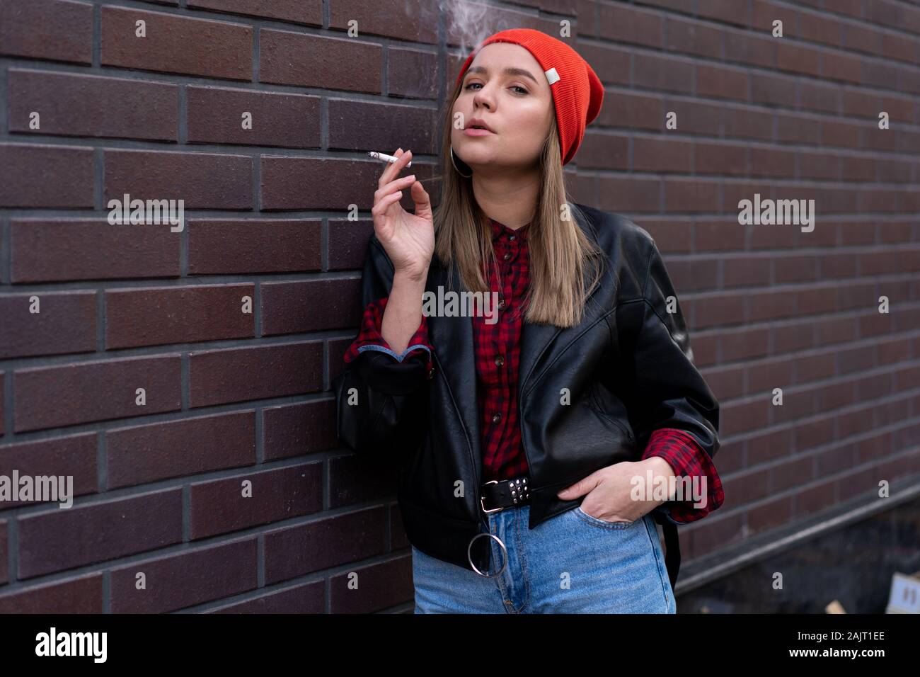 Beautiful young stylishly dressed Caucasian girl smokes cigarette on the street. Smoking bad habit Nicotine addiction Unhealthy lifestyle.  Modern wom Stock Photo