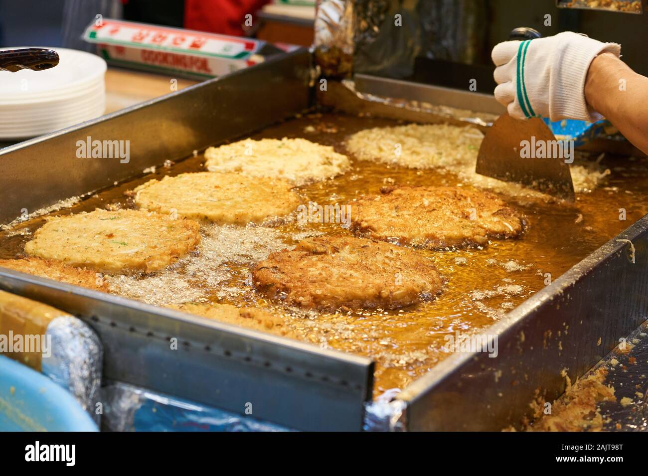 A cook uses a large metal spatula to move bindaetteok (bindae-tteok, mung bean pancakes) on a large oil-covered griddle at Gwangjang Market, Seoul. Stock Photo