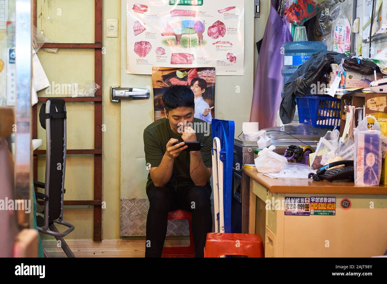 A Korean man in his 20s (twenties) watches a video on his smartphone while taking a break at his shop at Gwangjang Market in Seoul, South Korea. Stock Photo