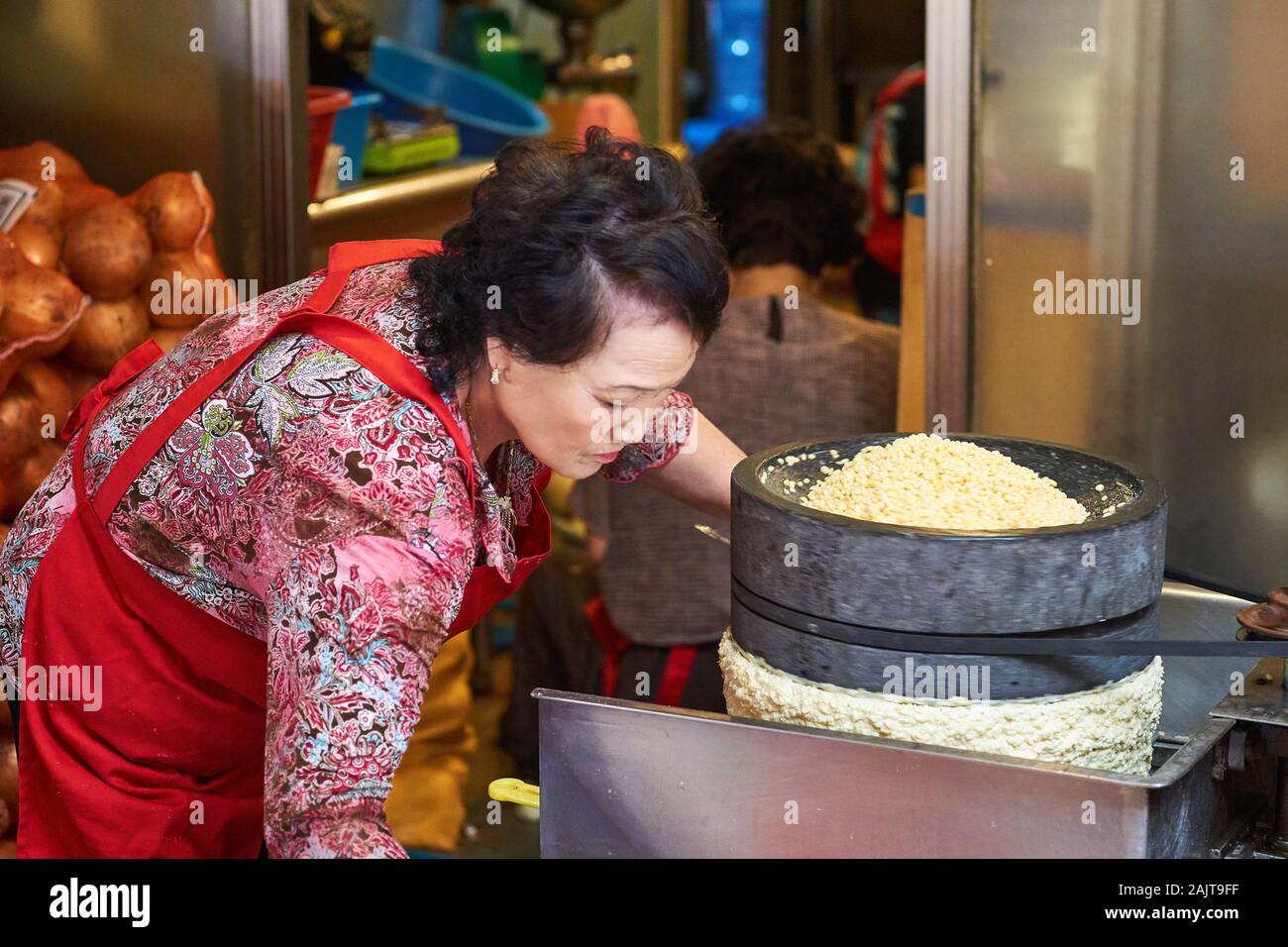 A Korean woman in red apron uses a special grinder to prepare mung beans for use in mung bean pancakes (bindae-tteok or bindaetteok) at Gwangjang Mkt. Stock Photo