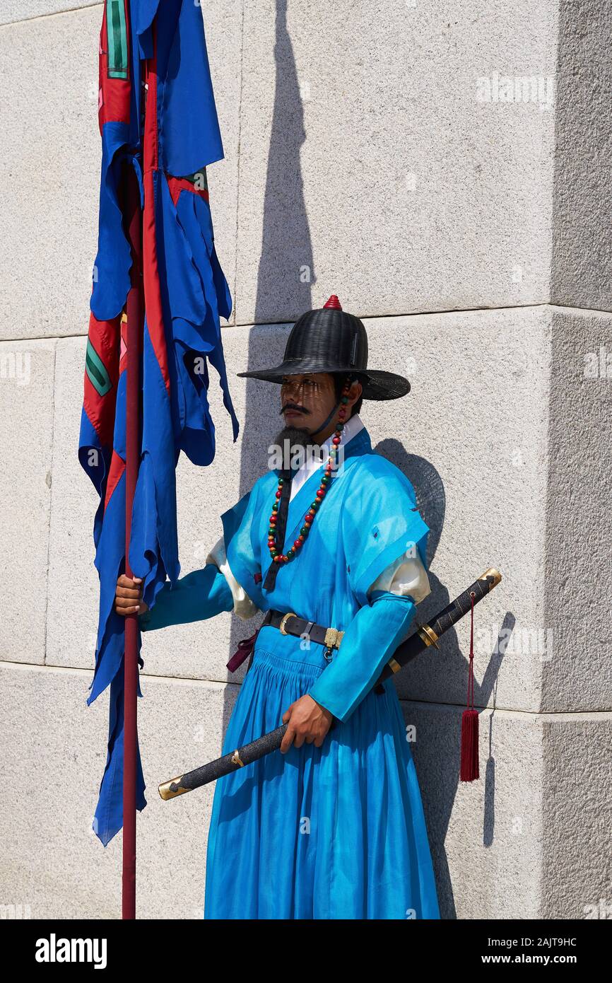 Royal Guard in blue Joseon era clothing bearing a standard stands before a gray stone wall at Gyongbokgung Palace. Stock Photo