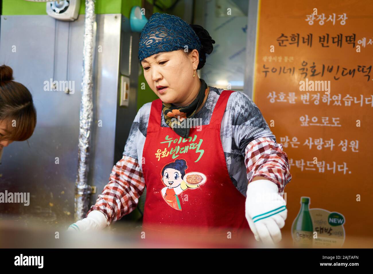 Female Korean chef cooks bindaetteok (bindae-tteok, mung bean pancakes) on a griddle at Gwangjang Market in Seoul, South Korea. Stock Photo
