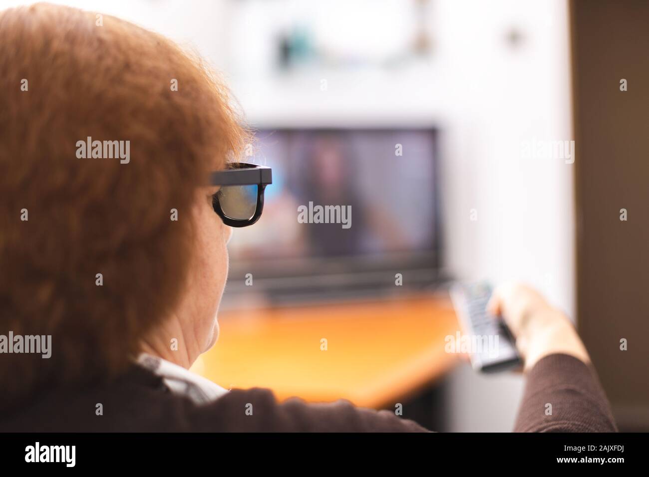 Senior lady with a 3d eyeglasses and tv remote point to a tv screen. Time to watching a tv. Remote control In hand. Focus on eyeglass Stock Photo