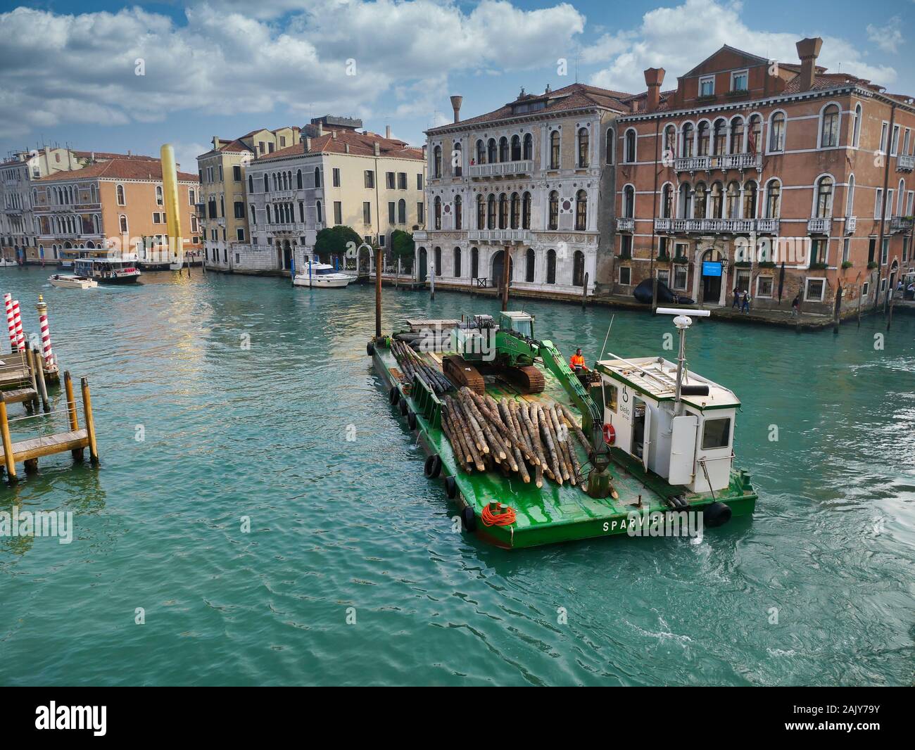 A maintenance barge carrying a supply of wooden pilings moves along a canal in central Venice, Italy on a cloudy day in Autumn. Stock Photo