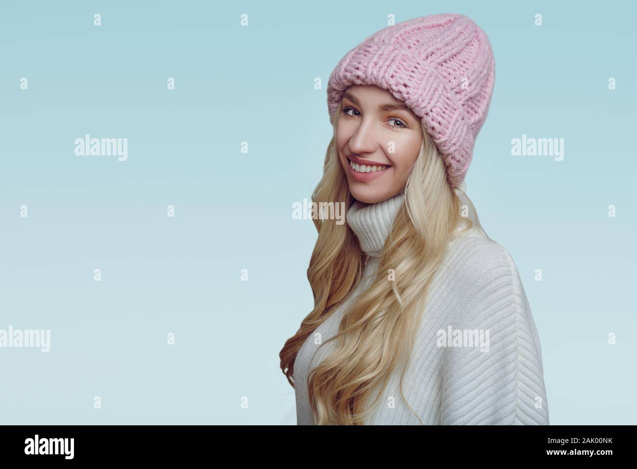 Happy young woman with long blond hair in pink knitted winter hat looking at the camera with a tourquoise background Stock Photo